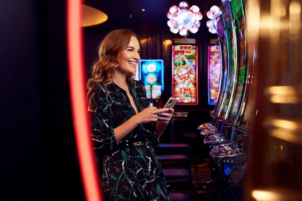 SPINARA Slots — Thousands of thrilling machines await A woman smiling by bright slot machines showing lucky symbols, showcasing the exciting slot offerings at SPINARA.